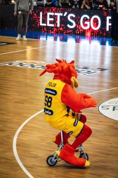Ein Maskottchen in einem bunten Kostüm fährt auf einem Roller über das Basketballfeld.