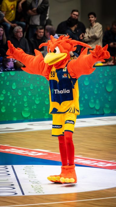 Ein fröhlicher Maskottchen mit orangem Gefieder und Sportkleidung feiert auf dem Basketballfeld.