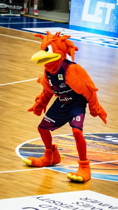 Ein orangefarbener Basketball-Maskottchen in sportlicher Pose auf dem Spielfeld.