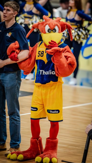 Maskottchen mit orangenem Fell in Basketballkleidung, umgeben von Cheerleadern.