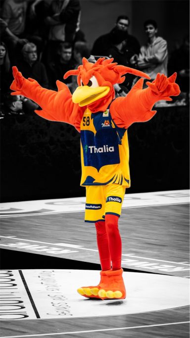 Ein orangefarbener Maskottchen mit Vogeldesign in Basketballkleidung auf dem Spielfeld.