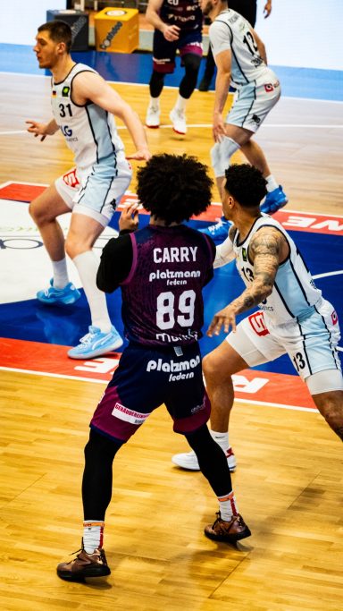 Basketballspiel mit Spielern in Trikots, einer spielt am Ball, andere verteidigen.