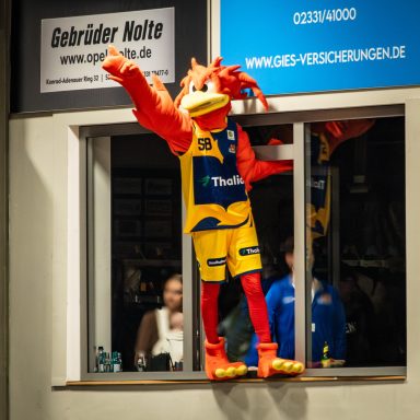 Ein bunter Maskottchen mit orangenen Haaren und Sportkleidung steht am Fenster.