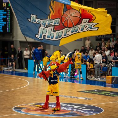 Ein Maskottchen in Rot und Gelb schwenkt eine große Basketballfahne in einer Sportarena.