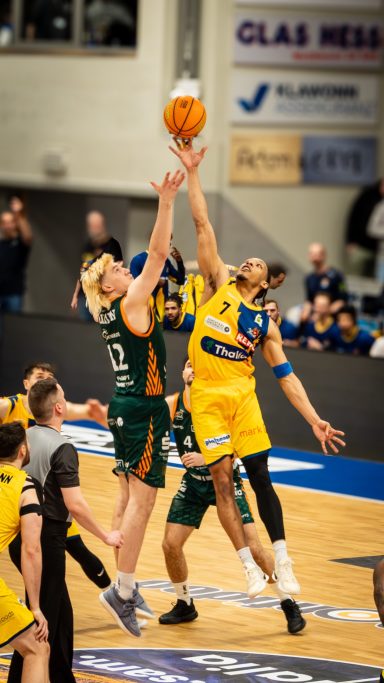 Zwei Basketballspieler springen um den Ball in einem mit Zuschauern gefüllten Stadion.